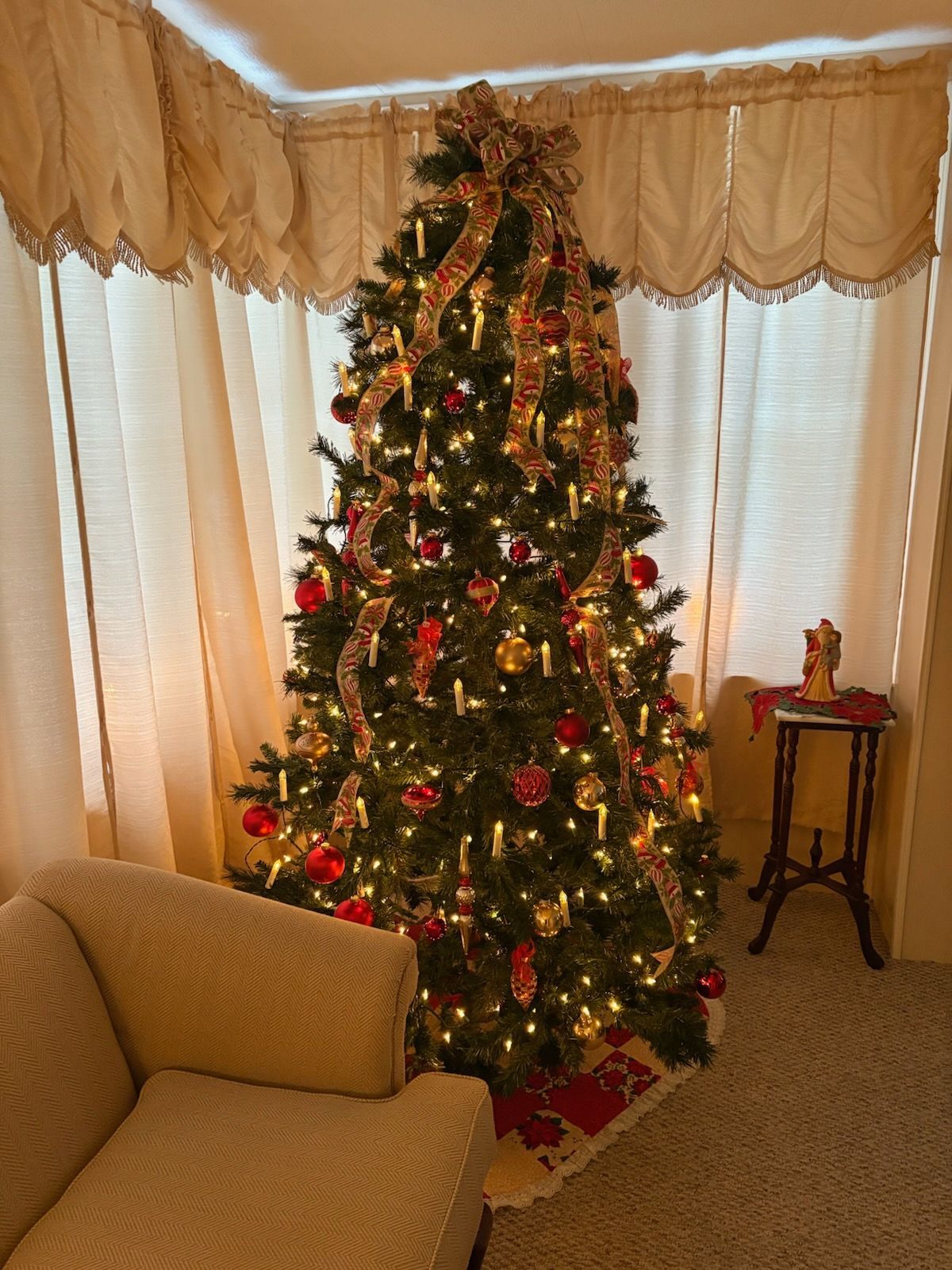 A Christmas tree decorated with red and gold ornaments and lights, positioned in a corner in front of beige curtains.