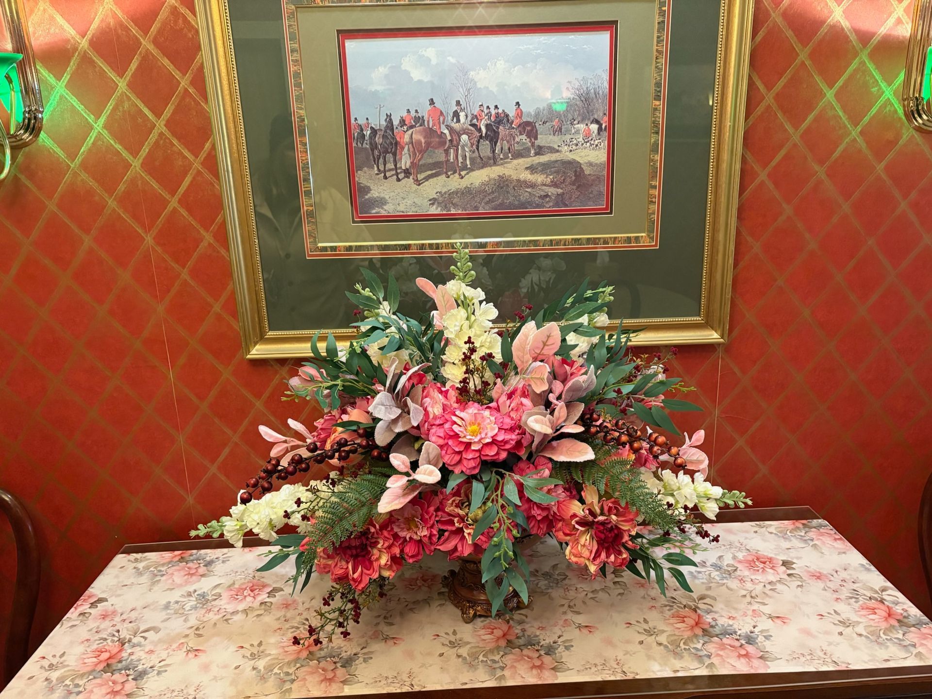 A large, colorful floral arrangement sits on a floral table runner below a framed hunting scene on a textured orange wall.