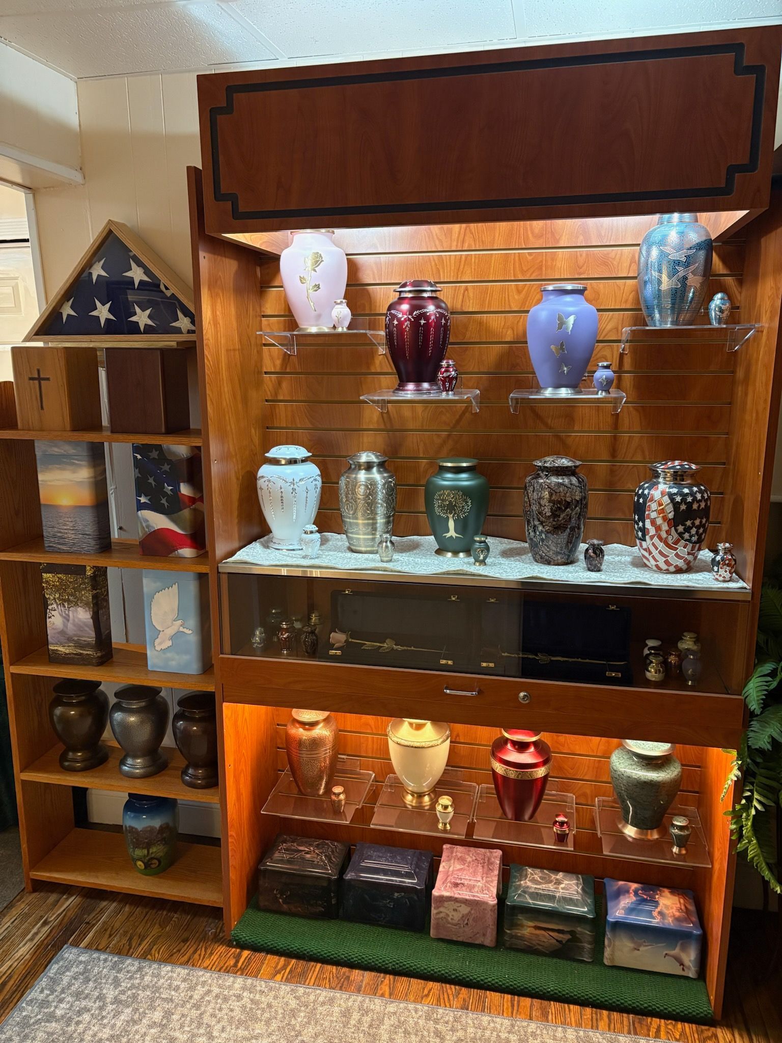 A wood shelving unit displays various funeral urns in a shop setting, with a flag case on a separate side shelf.