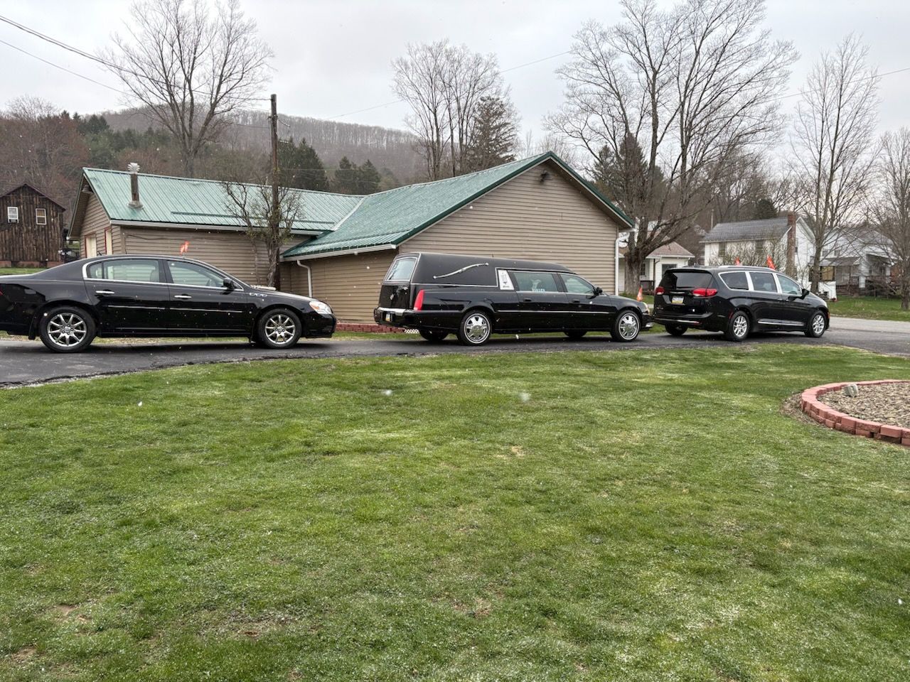 Three black funeral vehicles, including a hearse, parked in a row on a driveway in front of a house on a cloudy day.