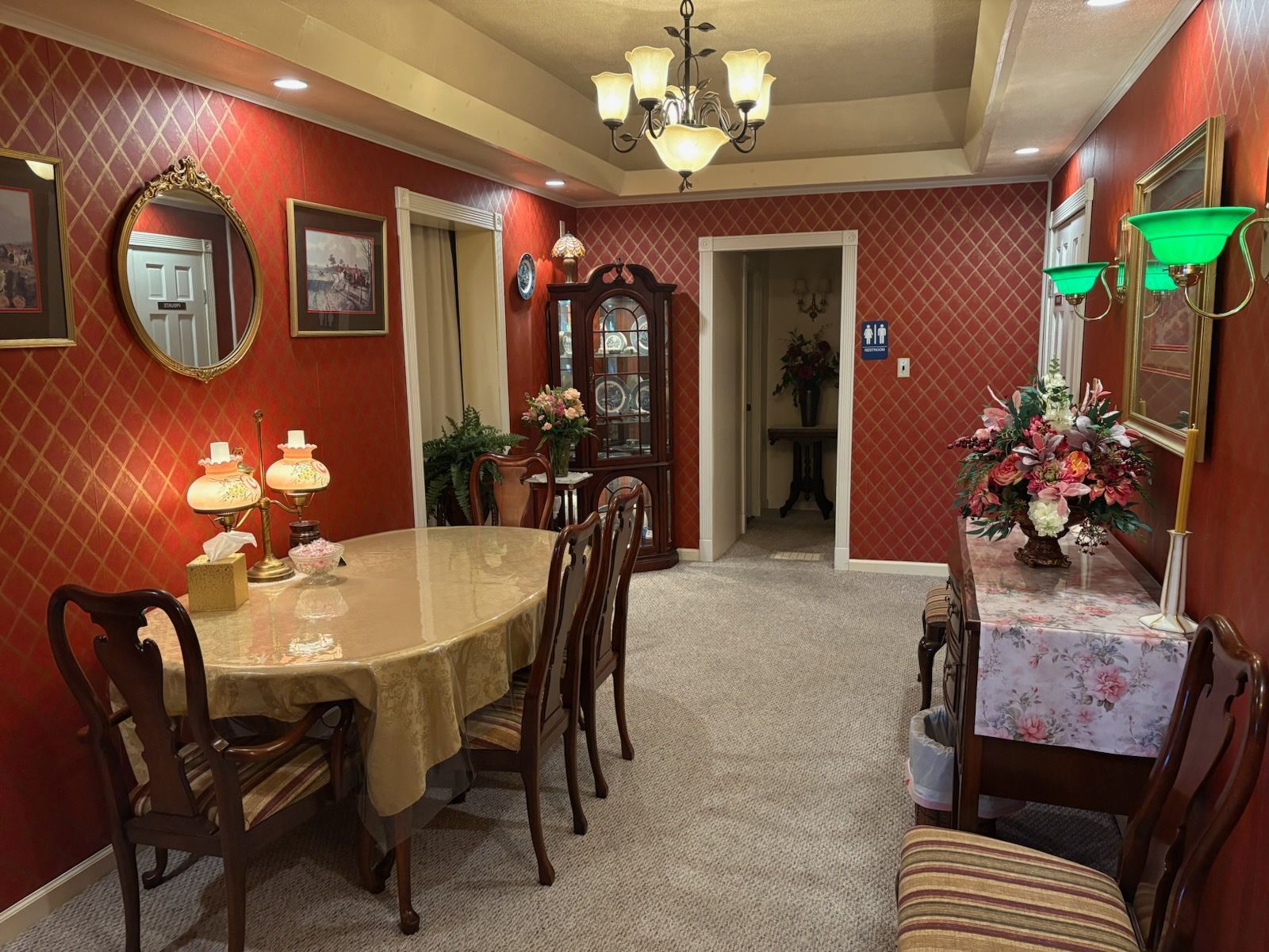 A dining room with red patterned wallpaper, an oval table with chairs, a glass-front cabinet, and a floral centerpiece.