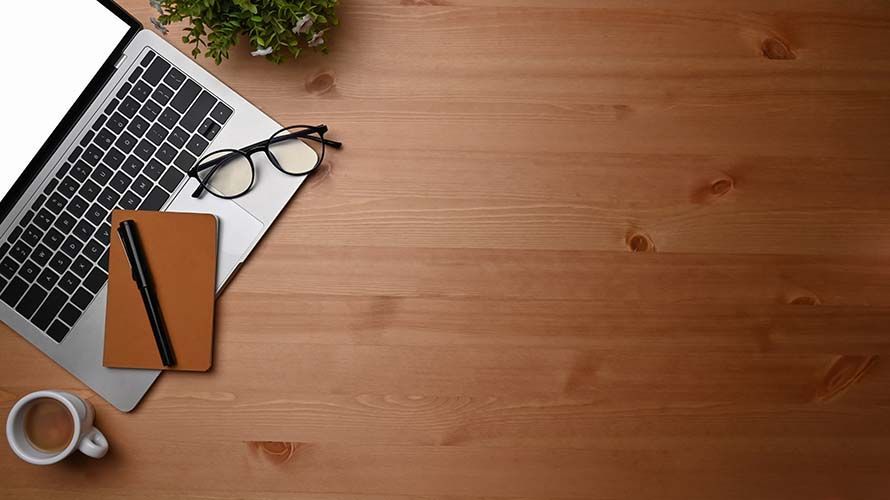 A laptop, a notebook with a pen, glasses, and a coffee mug sit on the corner of a wooden desk with a plant.