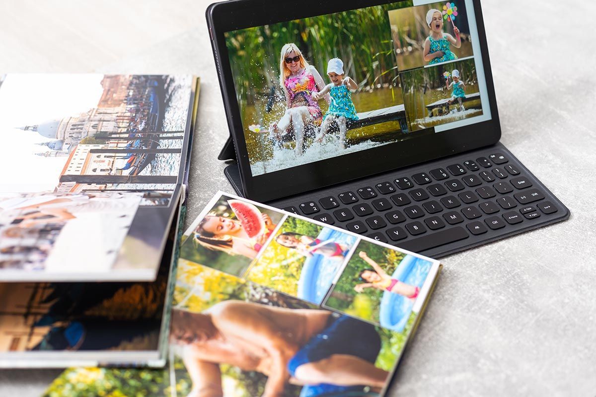 A tablet displaying photo collages sits next to open printed photo books on a light surface.