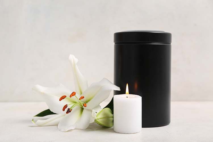 A black cremation urn sits next to a white lily and a lit white candle against a light background.
