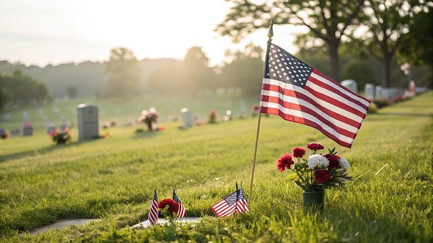 A U.S. flag flies above a small bouquet of red and white flowers at a sunny, grass-covered cemetery with headstones.
