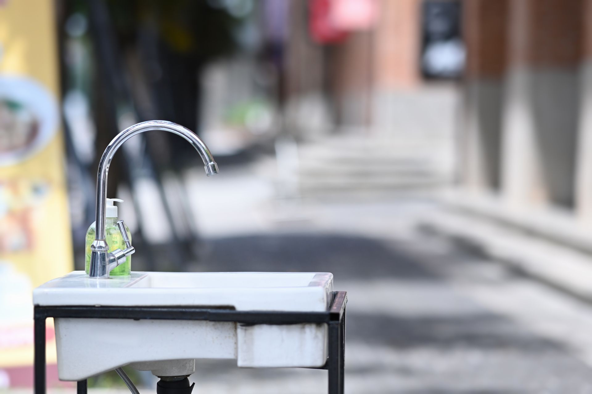 Small outdoor sink with a curved faucet and soap bottle on a table, against a blurred street background