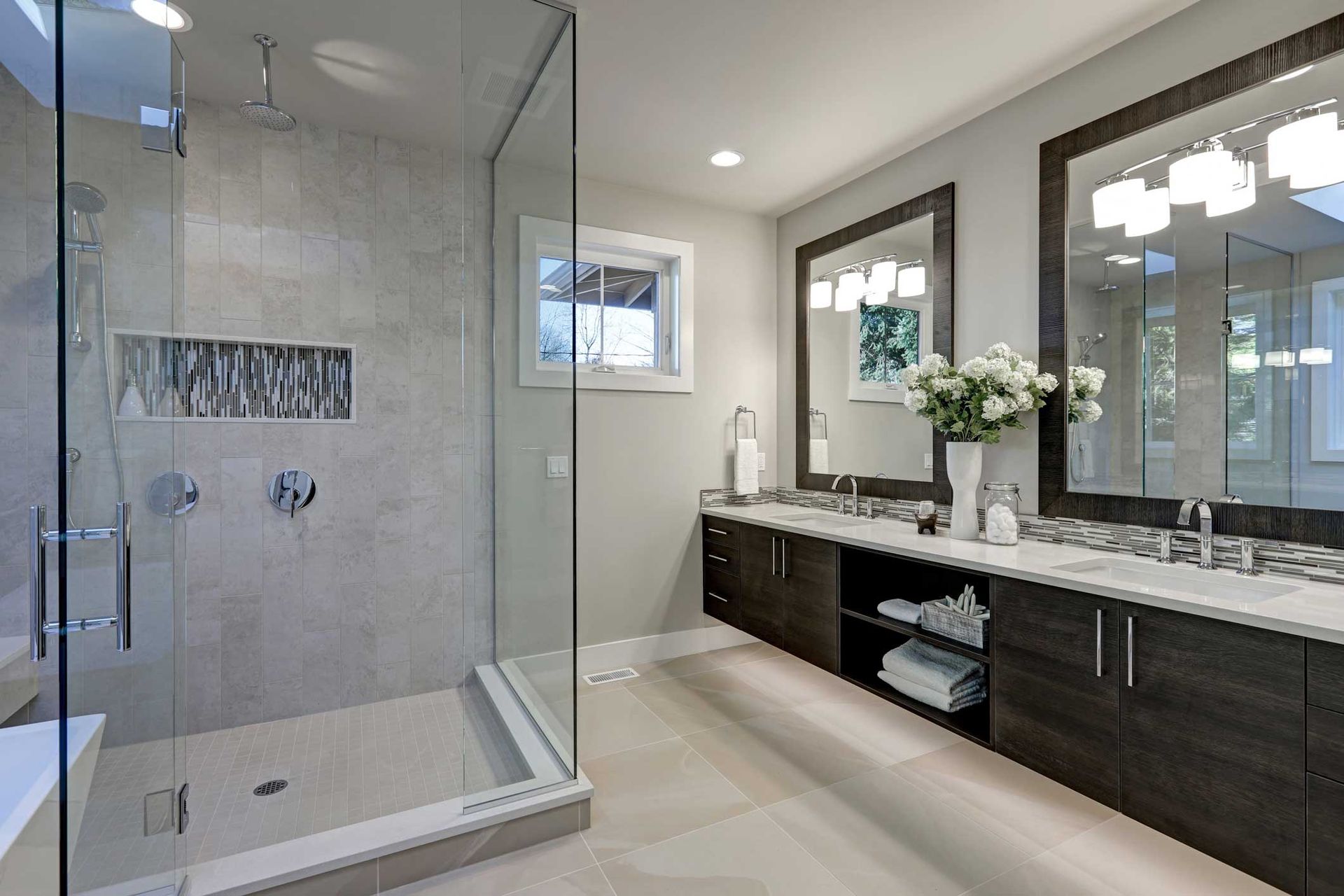 Modern bathroom with glass shower, dark cabinets, and dual sinks. Light gray walls, white countertops, and neutral floors.
