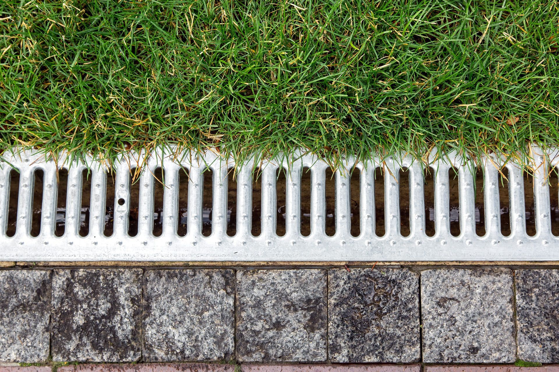 iron grate of a storm drainage system on the side of a footpath made of pavers near a green lawn top view