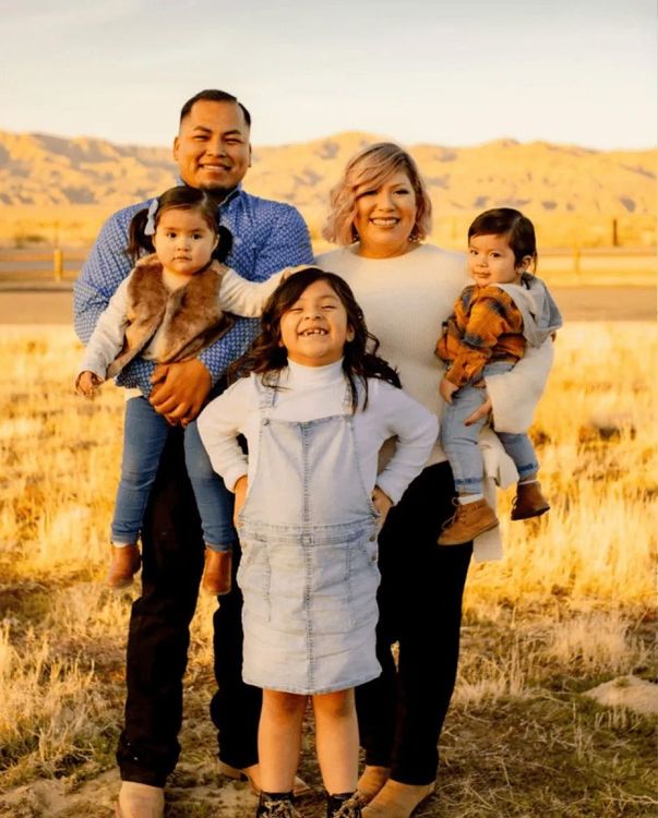 Family of five smiling outdoors: parents holding two babies, one child in front. Field, mountains.
