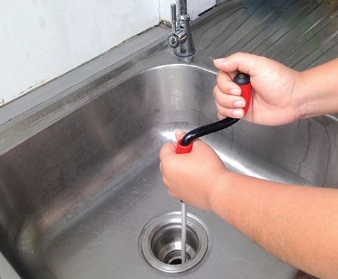 Hands use a drain snake to unclog a stainless steel kitchen sink.