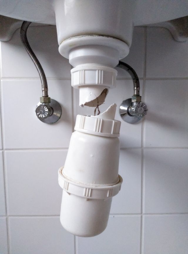 Broken white plastic P-trap under a sink, showing a cracked section and water supply lines.