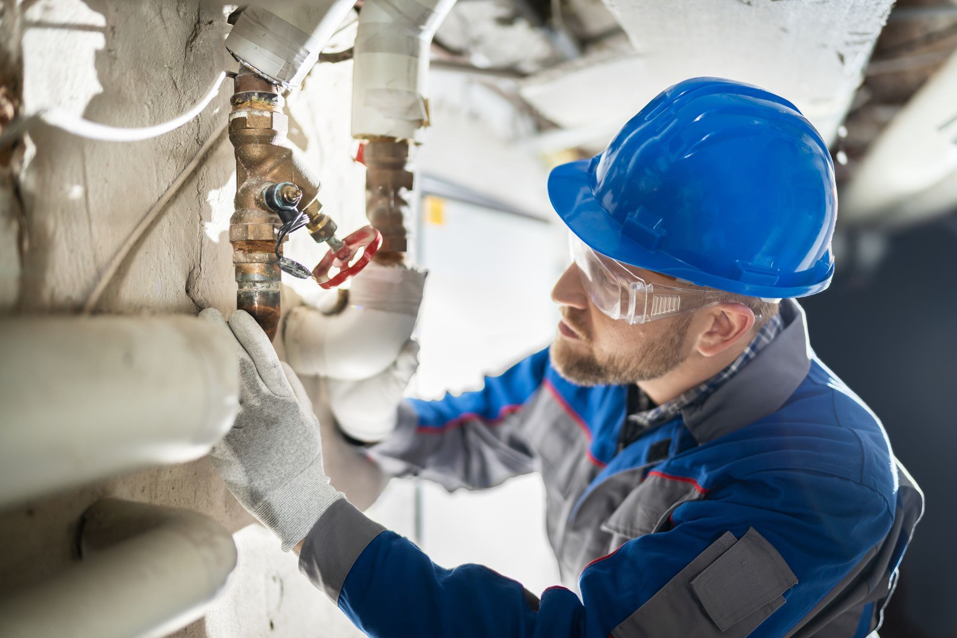 Plumber in blue hard hat examines pipes. Indoors, he wears safety glasses, gloves, and work uniform.