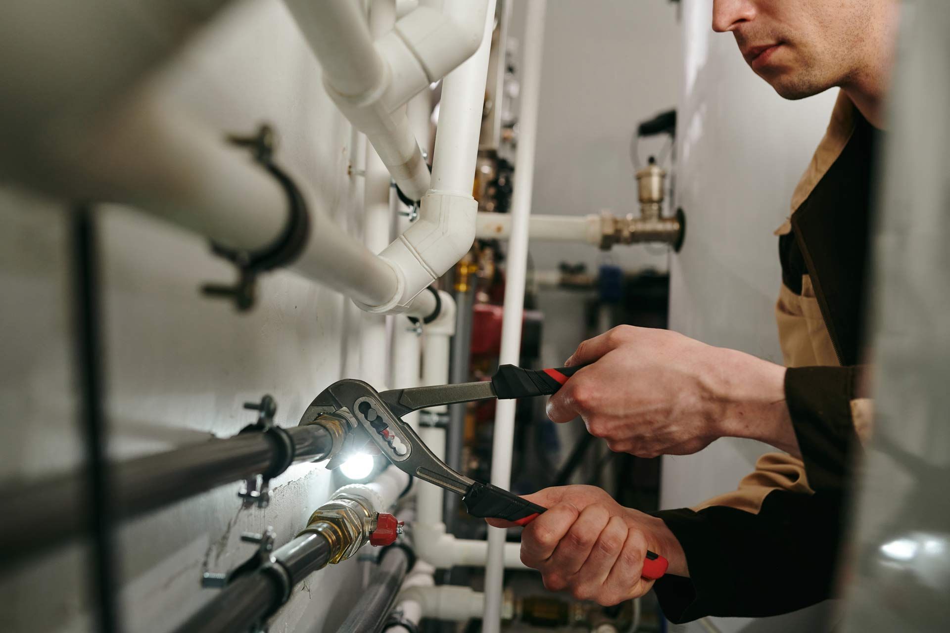 A commercial plumbing contractor using a tool to fix parts of pipes during work. Plumber using pliers to fix pipes in a mechanical room, illuminated by a flashlight.