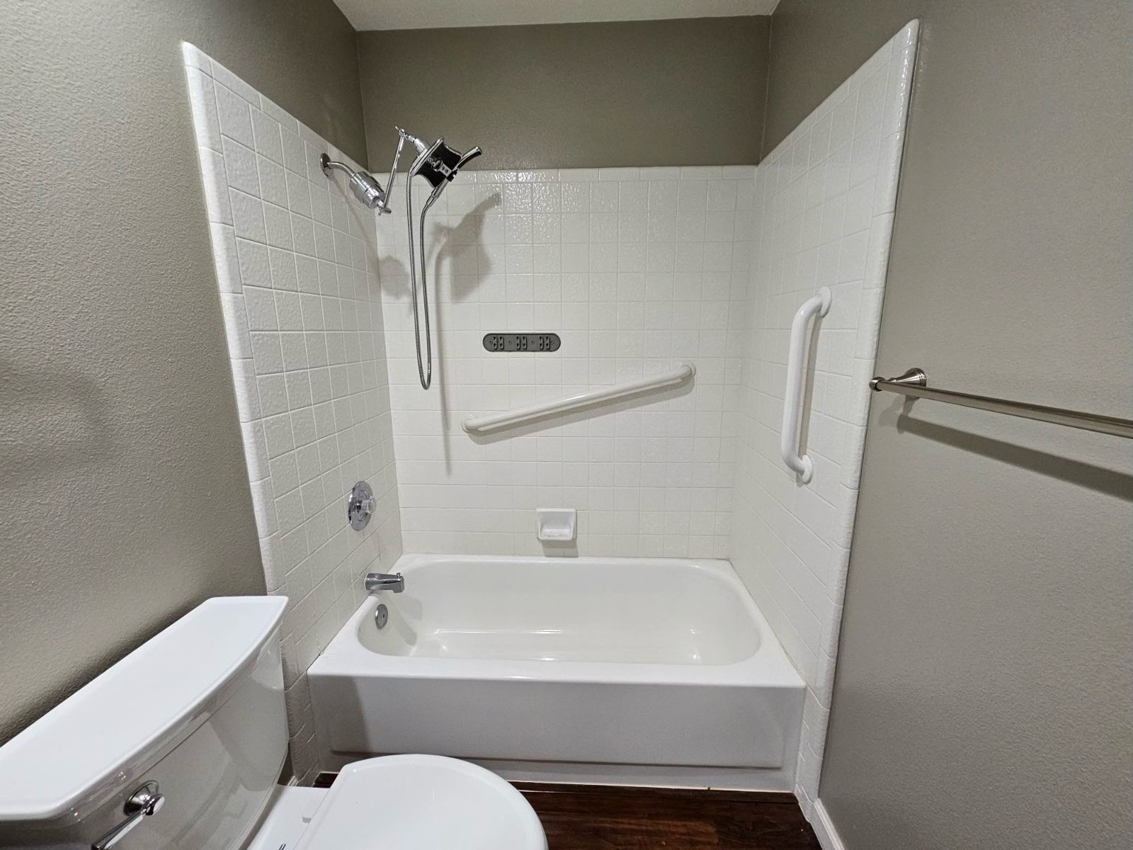 Small white bathtub and tiled shower with grab bars in a bathroom next to a toilet.
