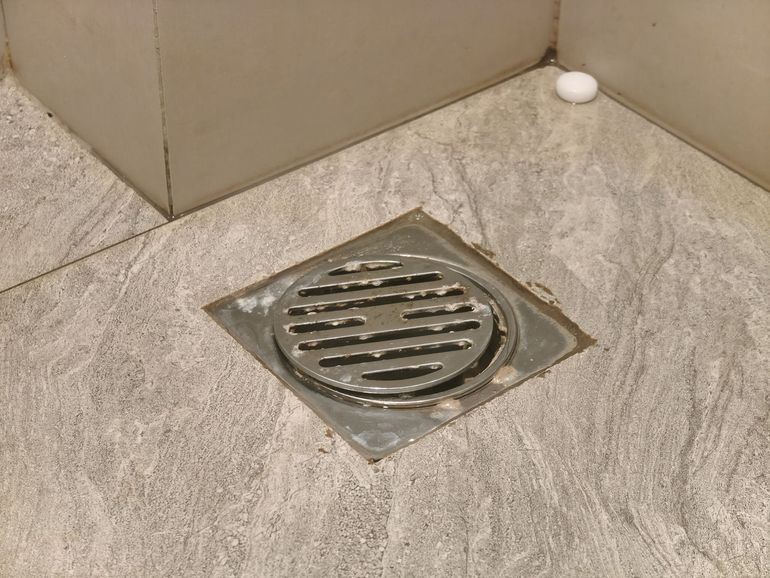 Close-up of a square floor drain in a gray tiled corner, with a white cap nearby.