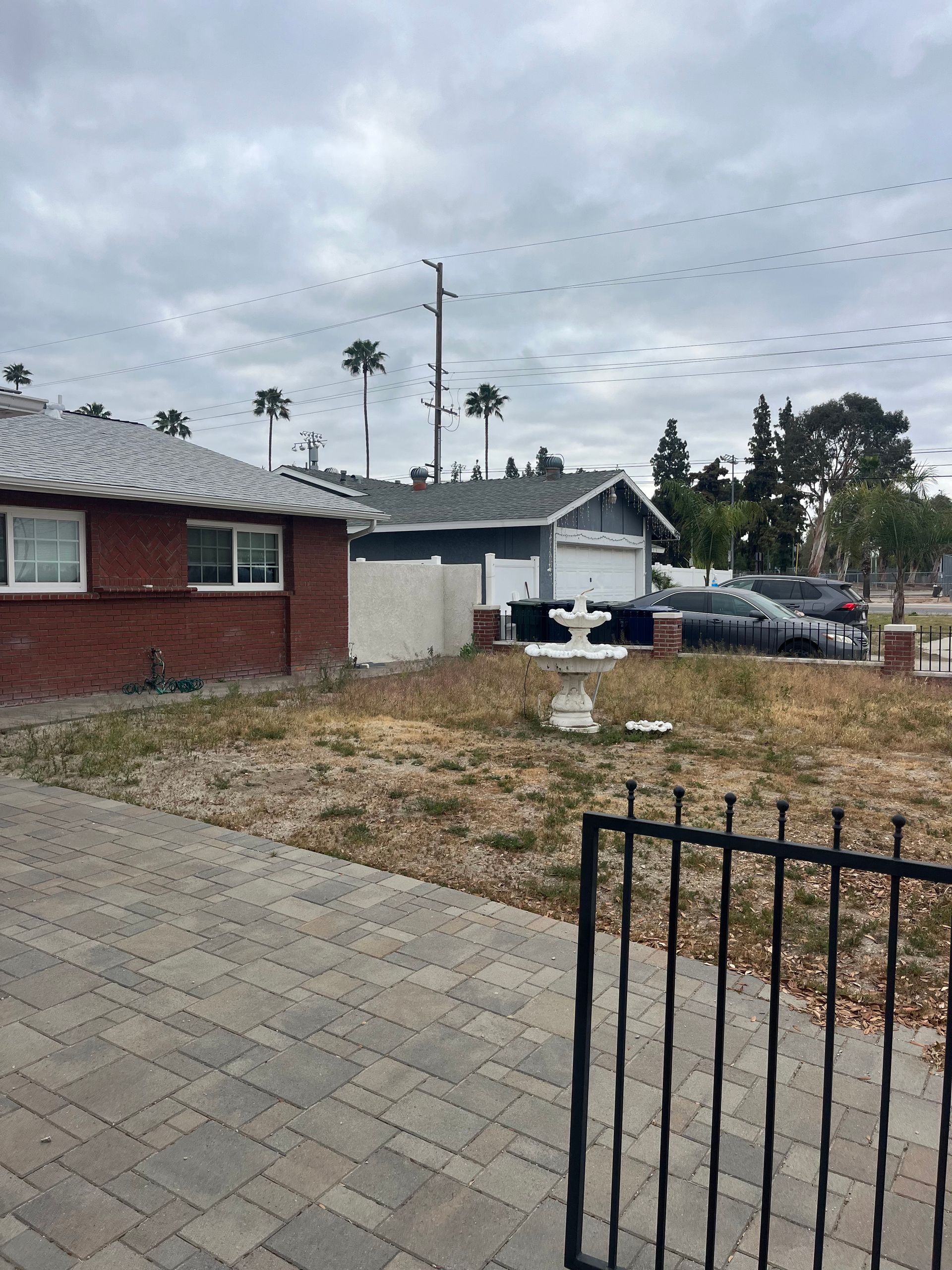 Suburban front yard with brick house, parked cars, and a black metal fence under cloudy skies