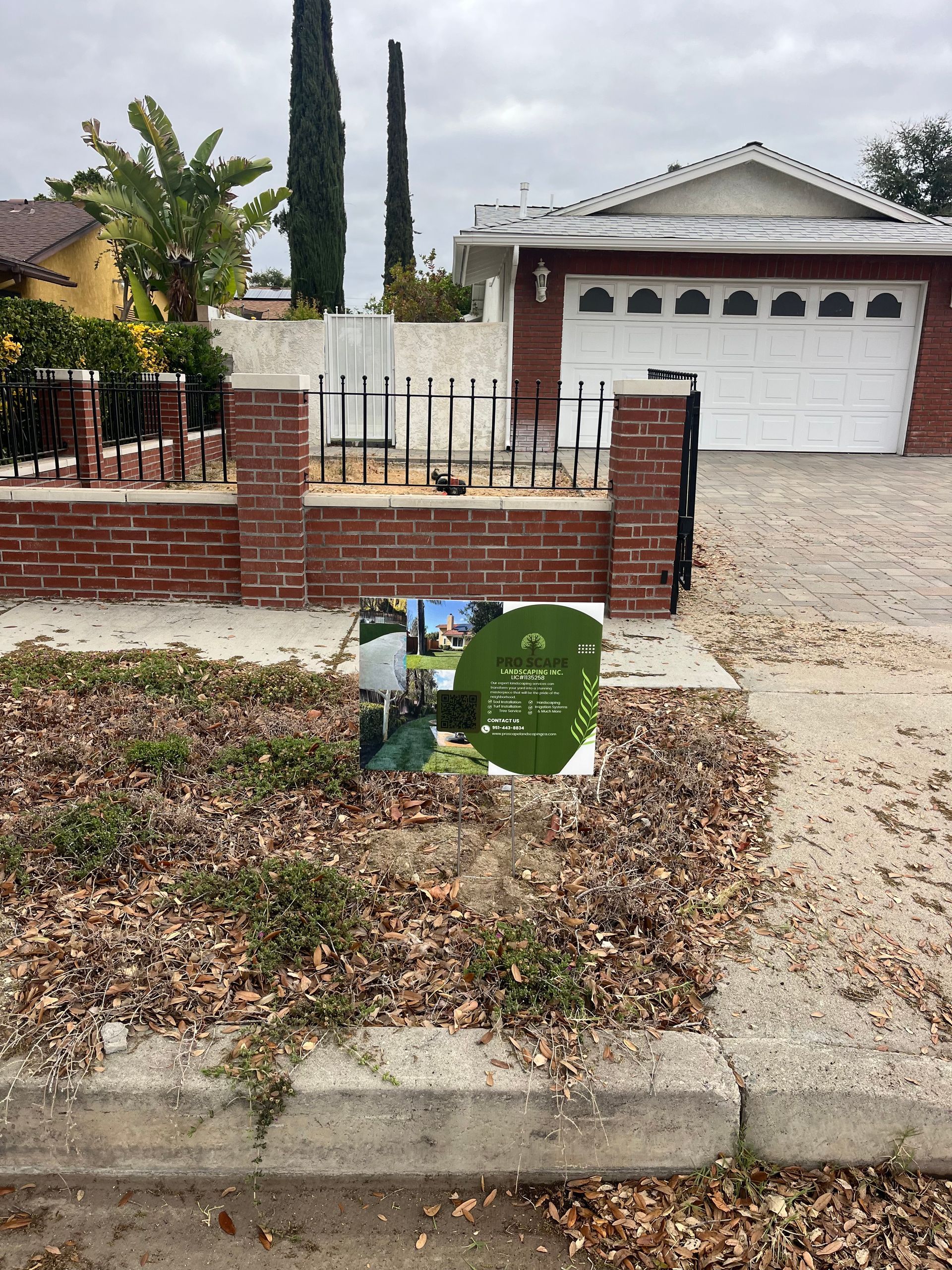 Front yard with brick wall, white garage, and a real estate sign near the curb.