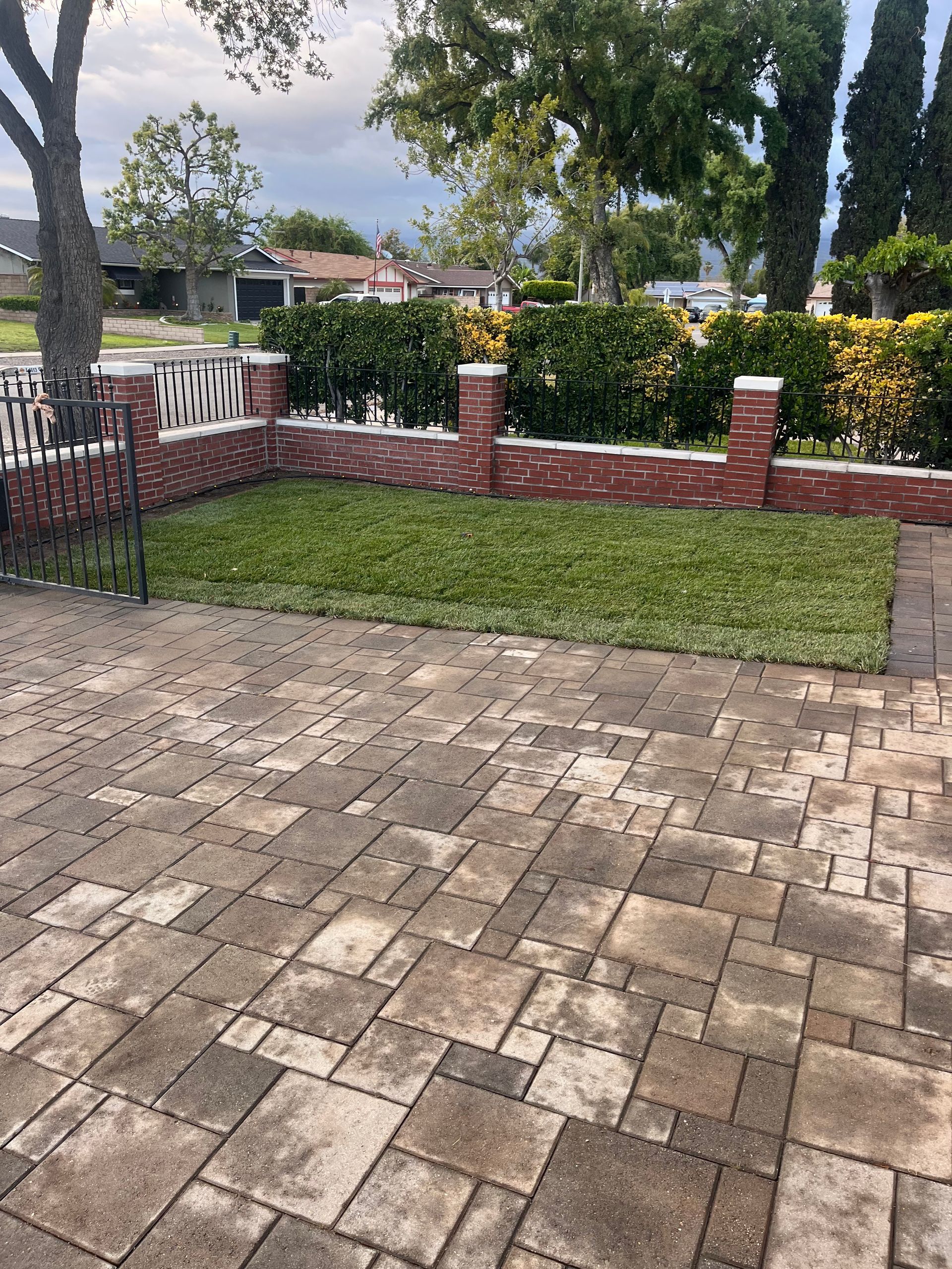Brick-paved patio overlooking a small fenced grass yard with trees and cloudy sky beyond