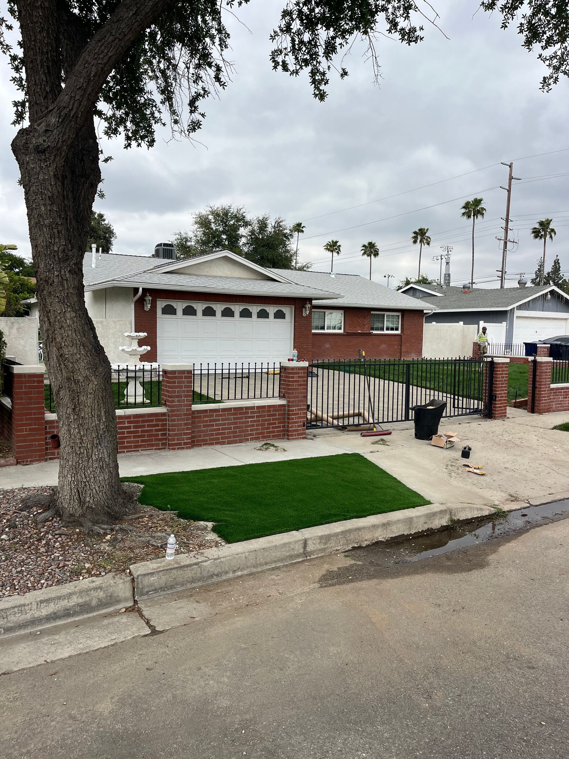 Single-story red-brick house with white garage, fenced front yard, and palm trees along a quiet street.