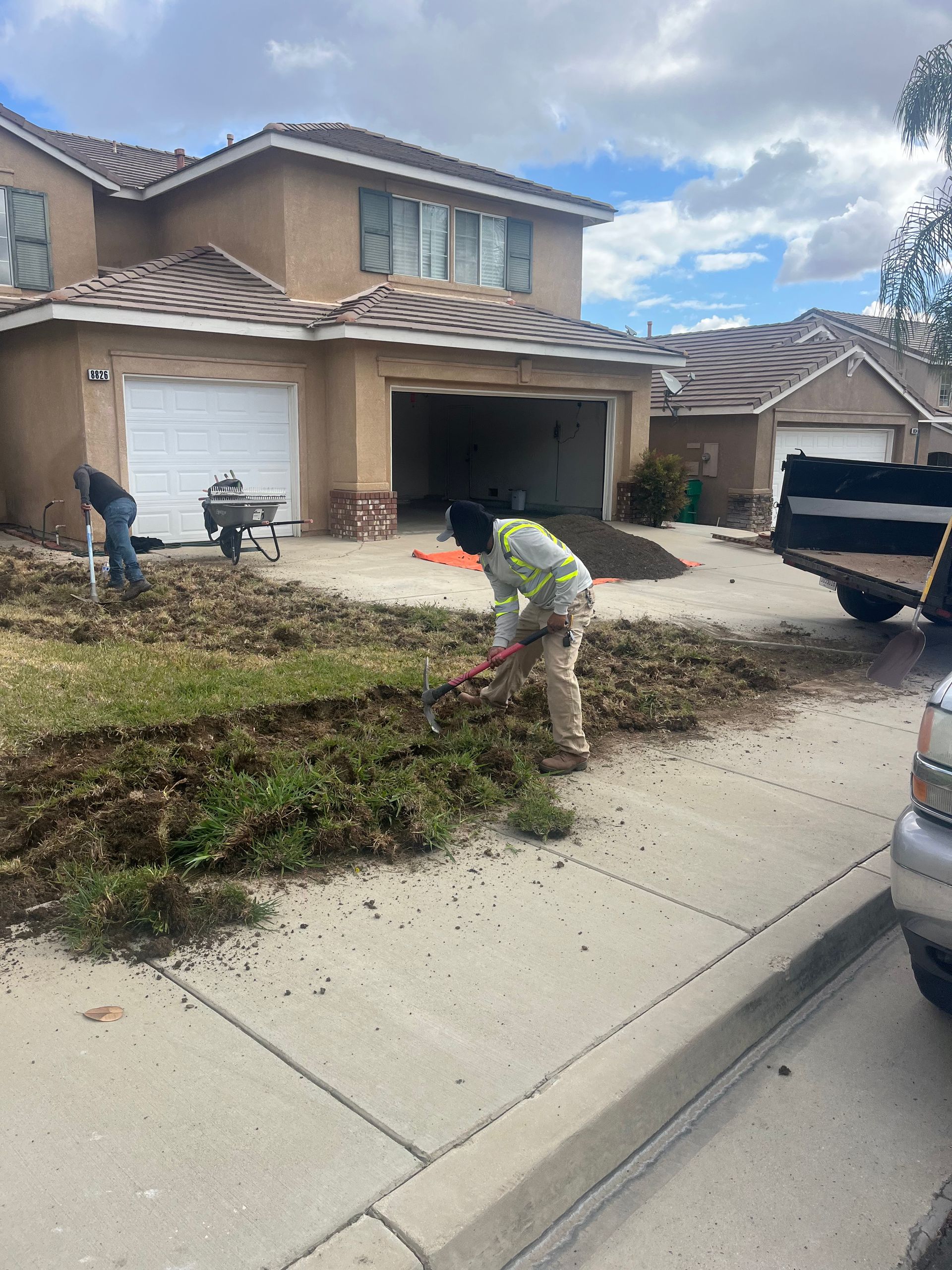 Worker raking dirt along a suburban front yard driveway beside a beige two-story house
