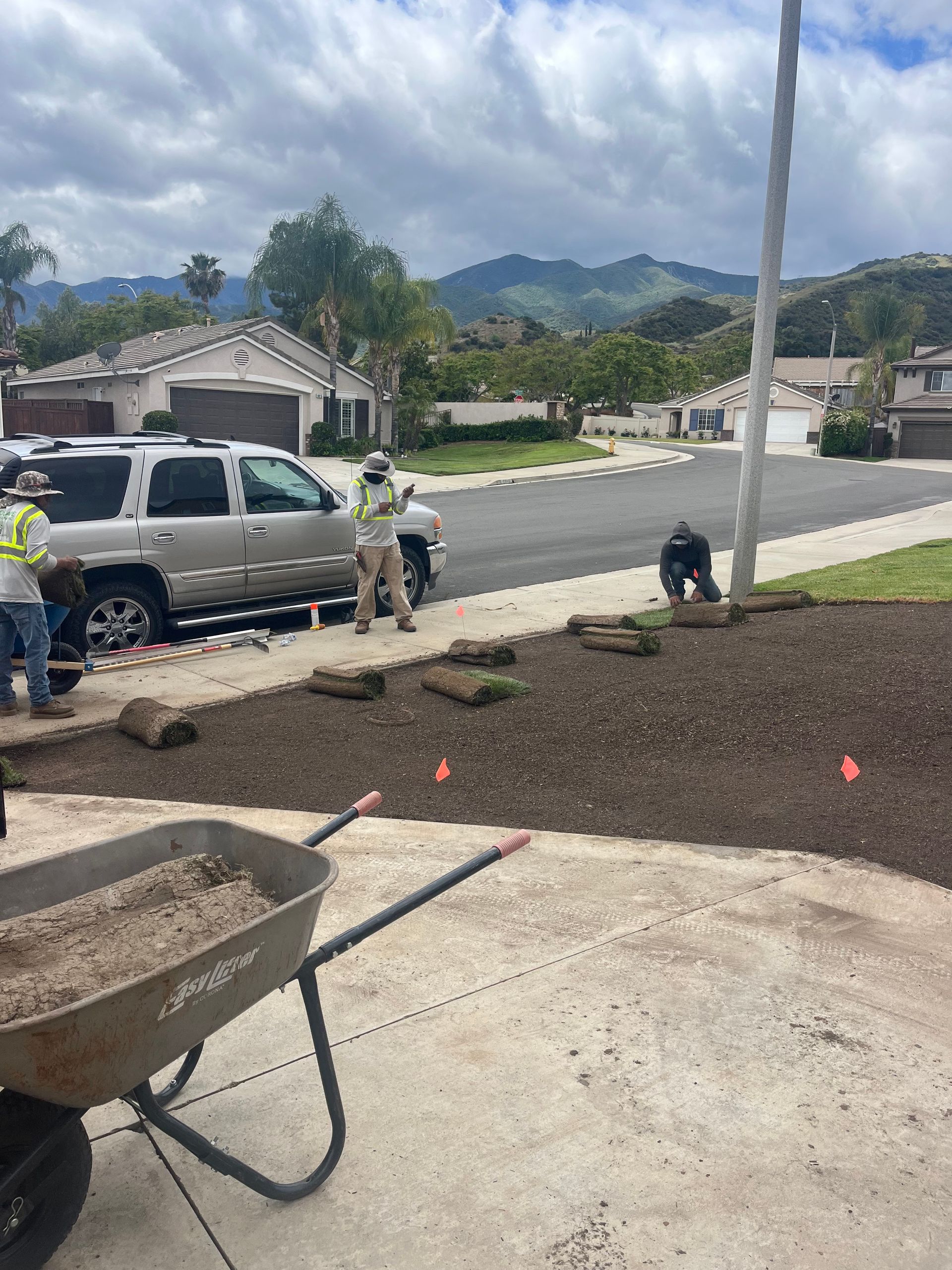 Roadwork on a suburban street with workers, a wheelbarrow, and fresh dirt beside parked vehicles and houses.