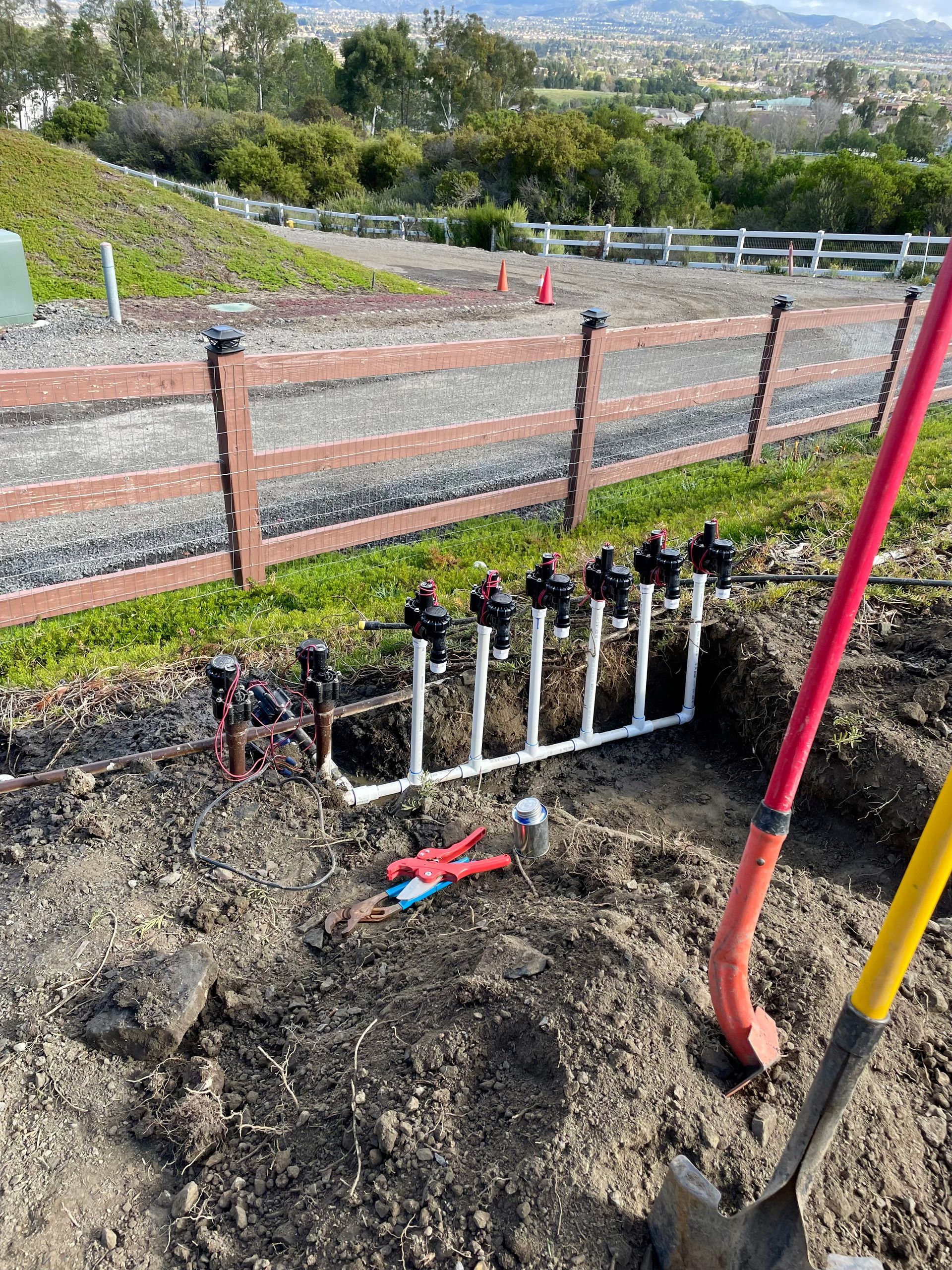 Irrigation system installation: PVC pipes with sprinklers, tools, shovel, and a brown fence in a dirt patch.
