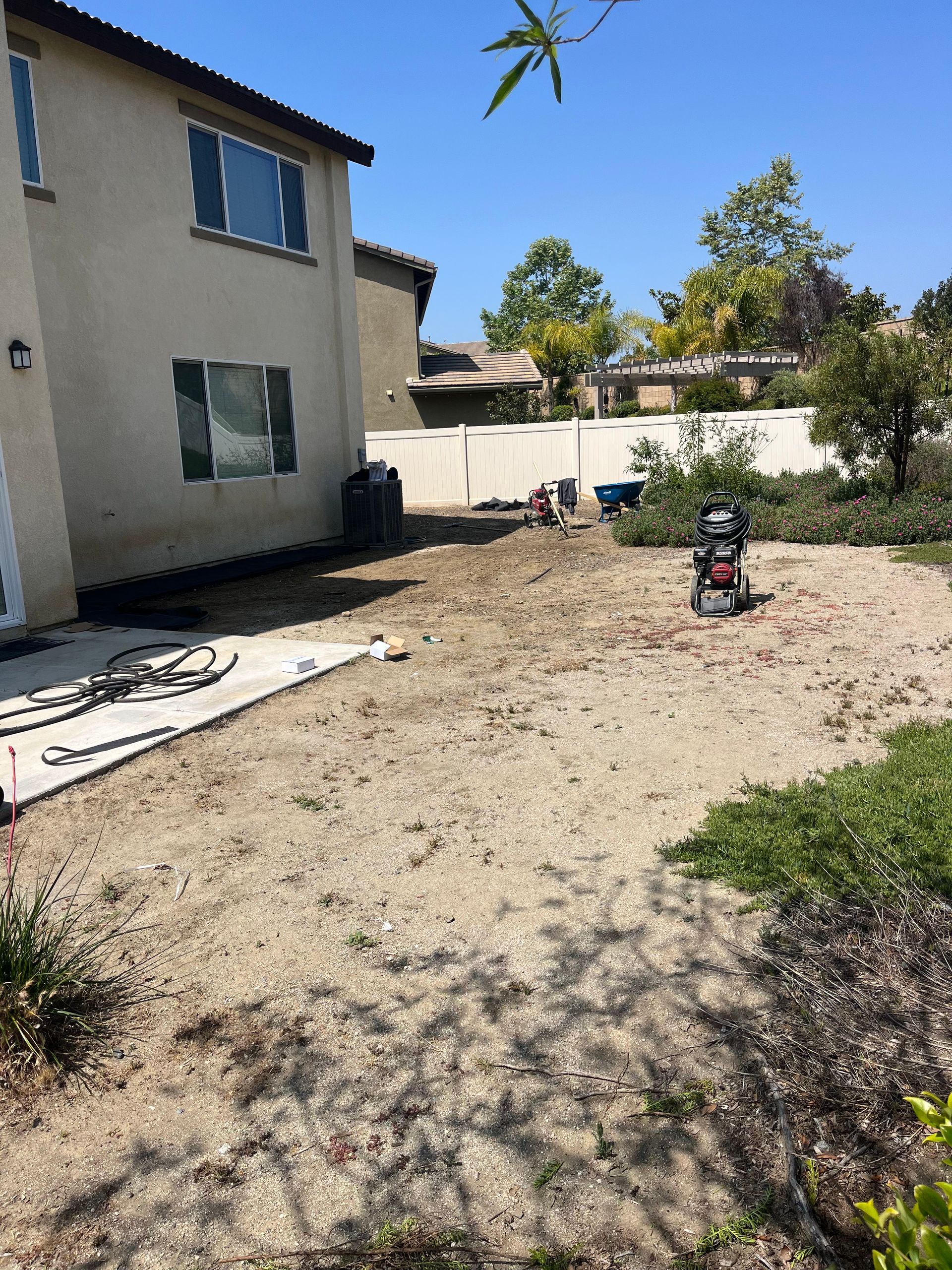 Backyard with dry dirt, a two-story house, white fence, and a small red lawn machine under a clear sky