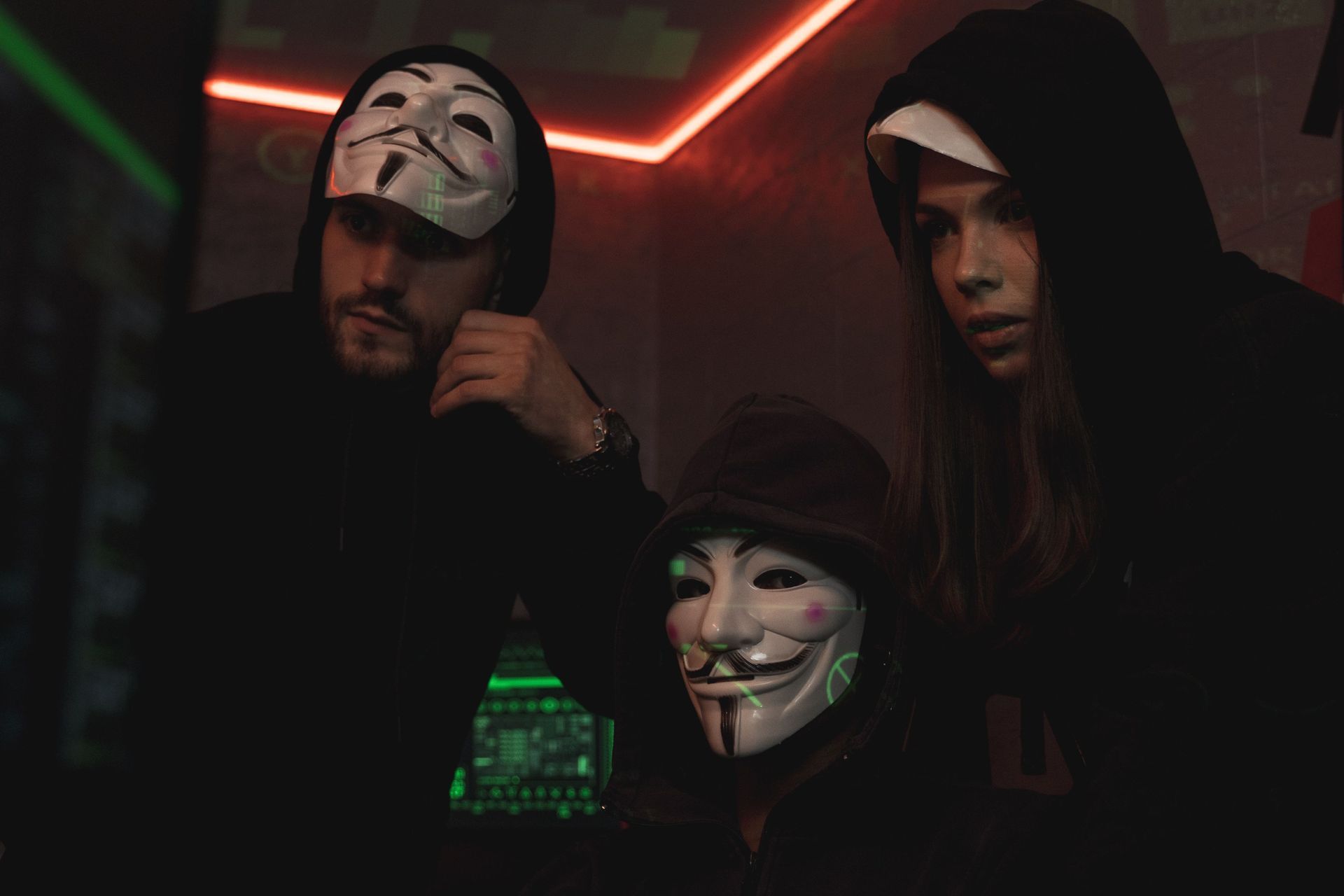 Three hooded people wearing Guy Fawkes masks in a dark neon-lit room