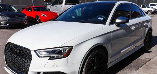 White Audi A3 with black accents parked on asphalt. Other cars are visible in the background.