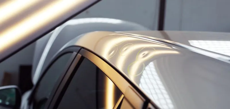 A close-up view of a car roof showing multiple small dents highlighted by a reflection board used for repair.