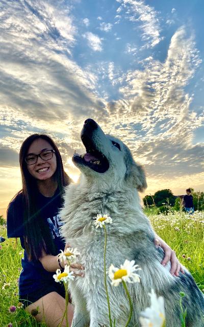 Woman with glasses smiles next to a large, light-colored dog in a field of wildflowers under a cloudy sky.