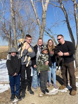 Family outdoors with two puppies; sunny day, trees in background.