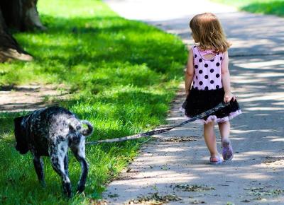 Girl in polka dot dress walks dog on a leash along a path next to green grass.