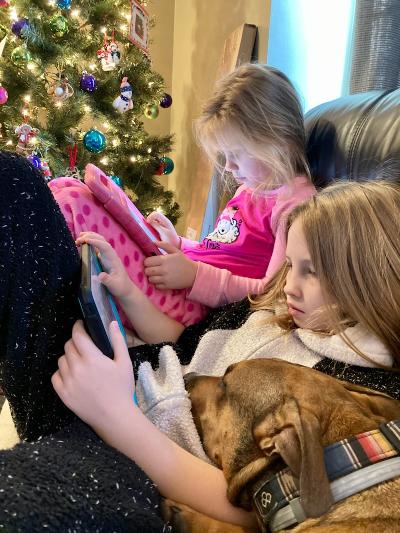 Two children using tablets next to a dog on a couch, Christmas tree in background.