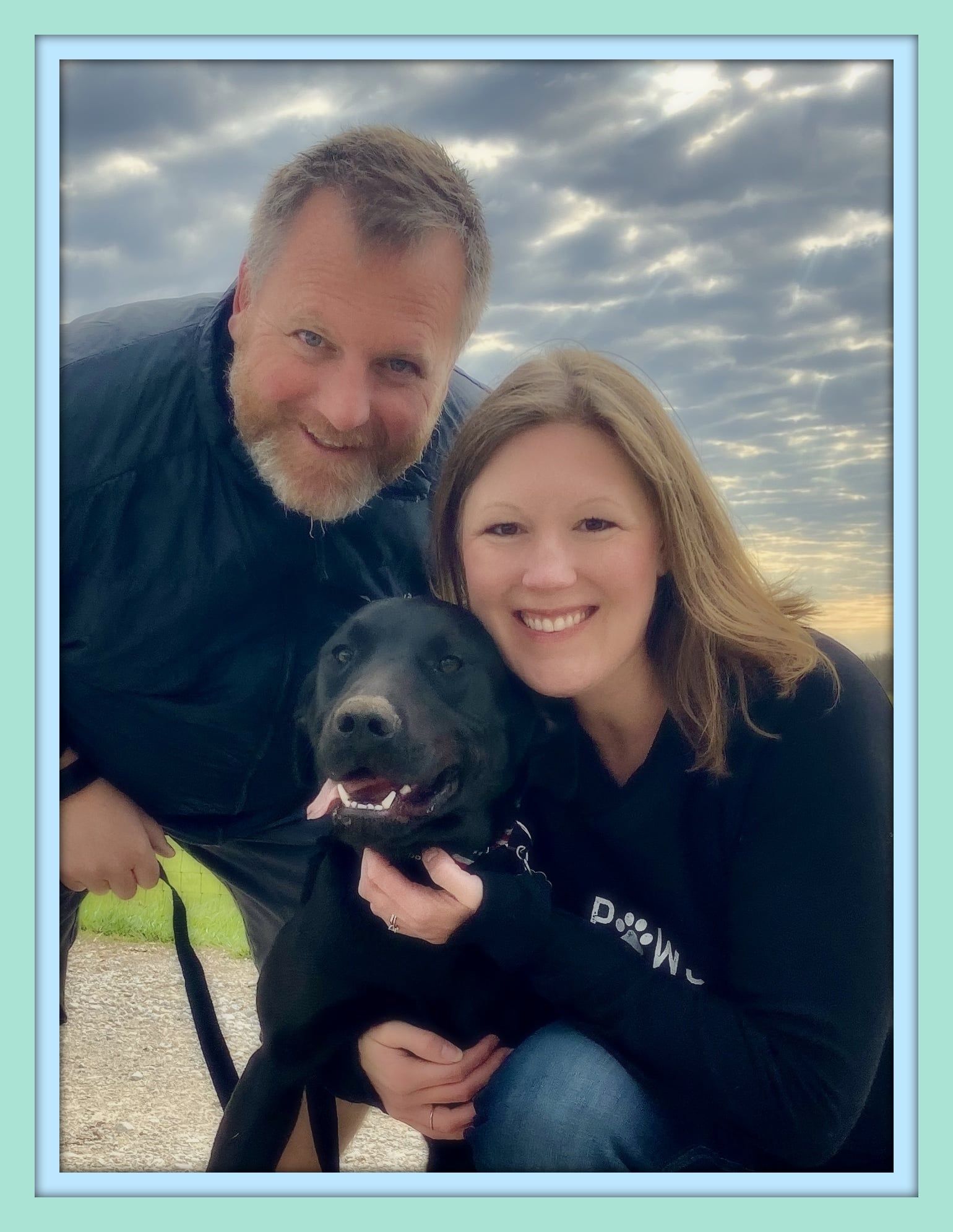 Couple smiling, cuddling a black dog outside under a cloudy sky.