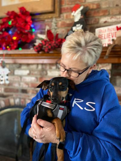 Woman in blue sweatshirt kisses small dog indoors near a fireplace with holiday decorations.