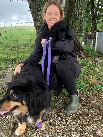 Woman holding a black dog and smiling, with a larger dog on leash in front of her. Outdoors on a cloudy day.