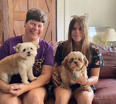 Two women smile, holding small dogs on a brown couch.
