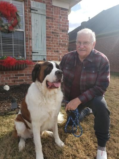 Man kneels next to a seated Saint Bernard dog outdoors. They both smile. Brick house in the background.