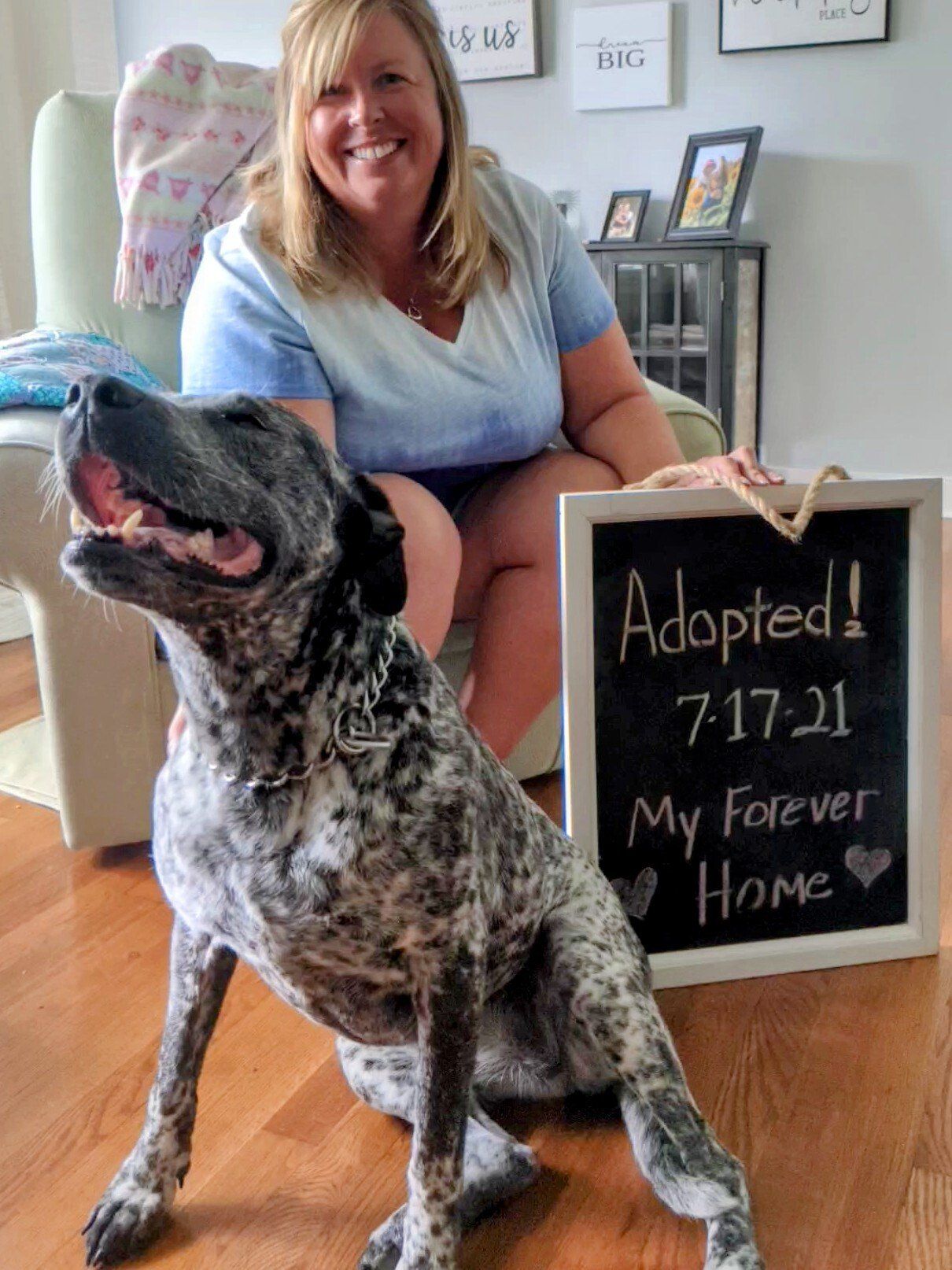 Woman sits with dog, both smiling. Dog's adoption date written on a chalkboard: 7-17-21, My Forever Home.