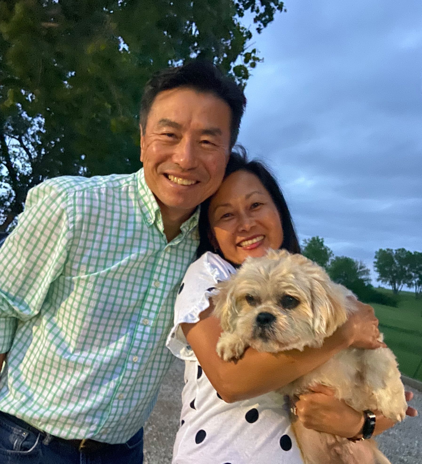 Couple smiles, one holding small dog, outside with trees and cloudy sky.