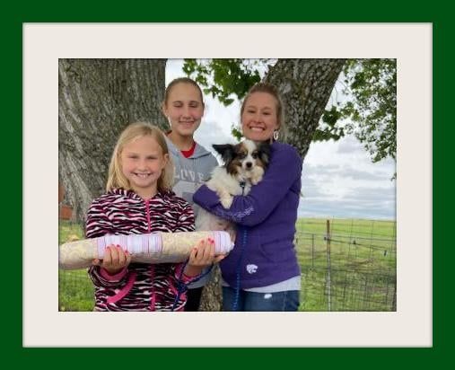Three smiling people with a small dog and a decorated corn husk in a green field.