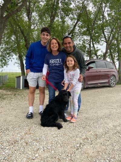 Family poses with a black dog in front of a car and trees.