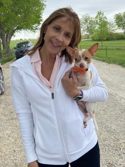 Woman in white jacket holds small dog wearing a bow tie, smiles outdoors.