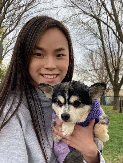 Woman smiling, holding small dog in purple sweater outside on a green lawn.