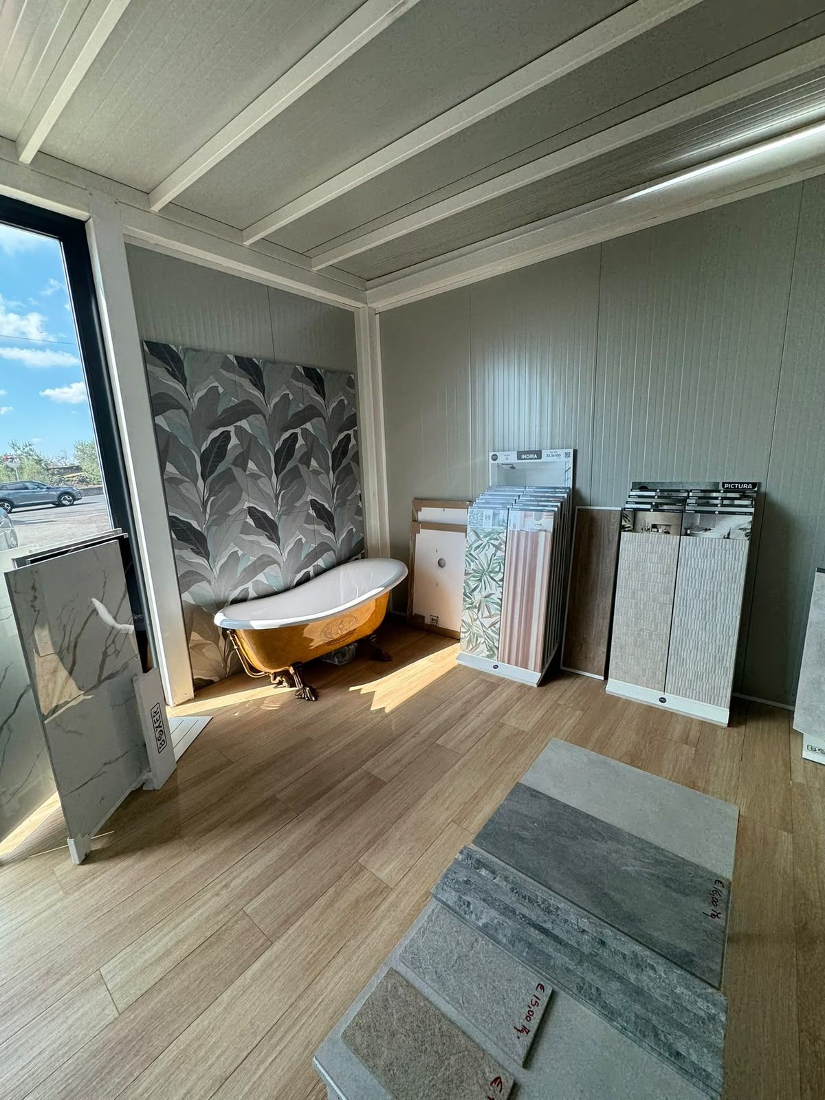Stanza moderna con vasca freestanding, pannello murale decorato e campioni di piastrelle accanto a una grande finestra.