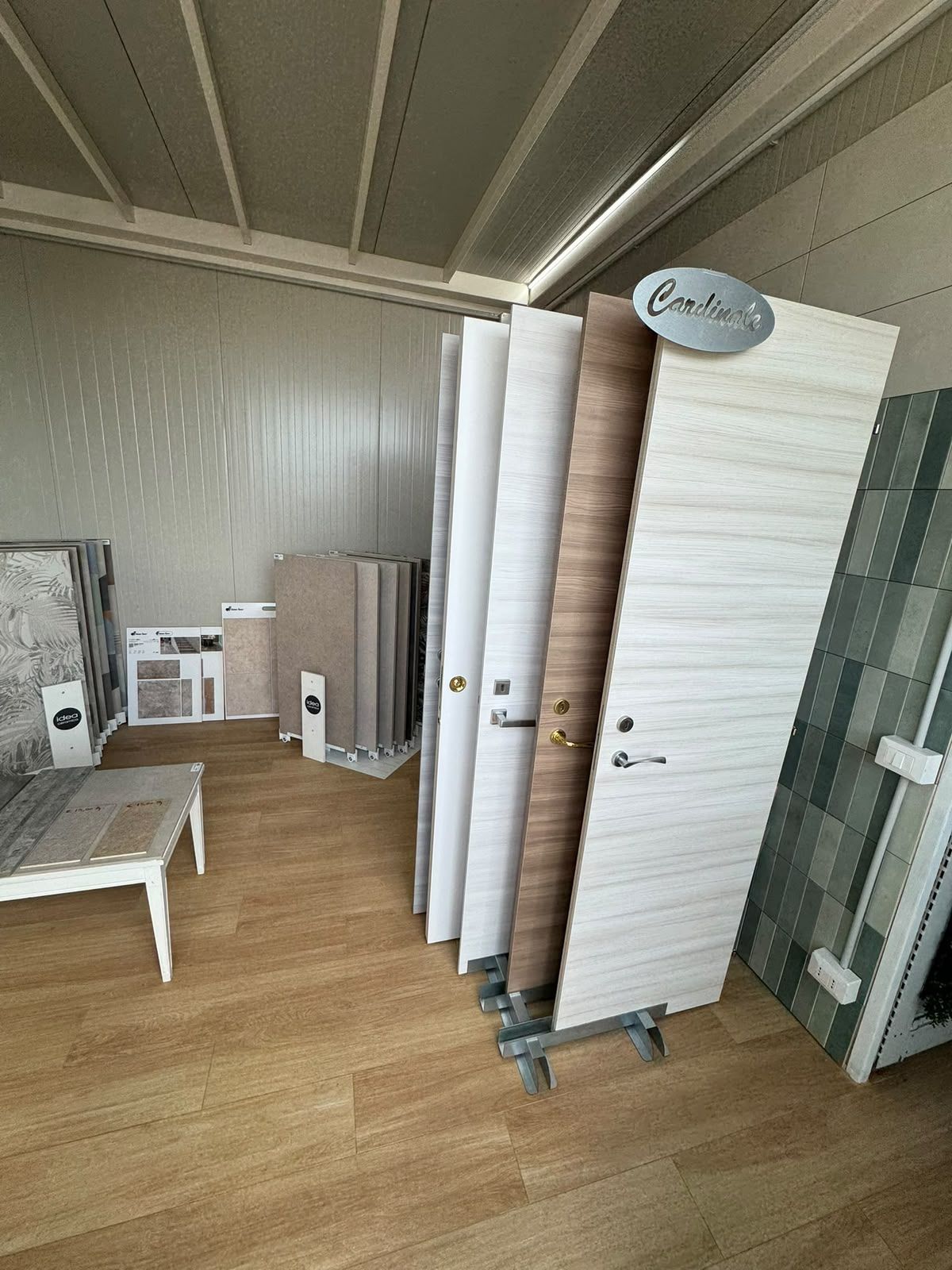 Showroom con pannelli campione impilati, una panca bianca e pannelli espositivi su un pavimento in legno, sotto un soffitto spiovente