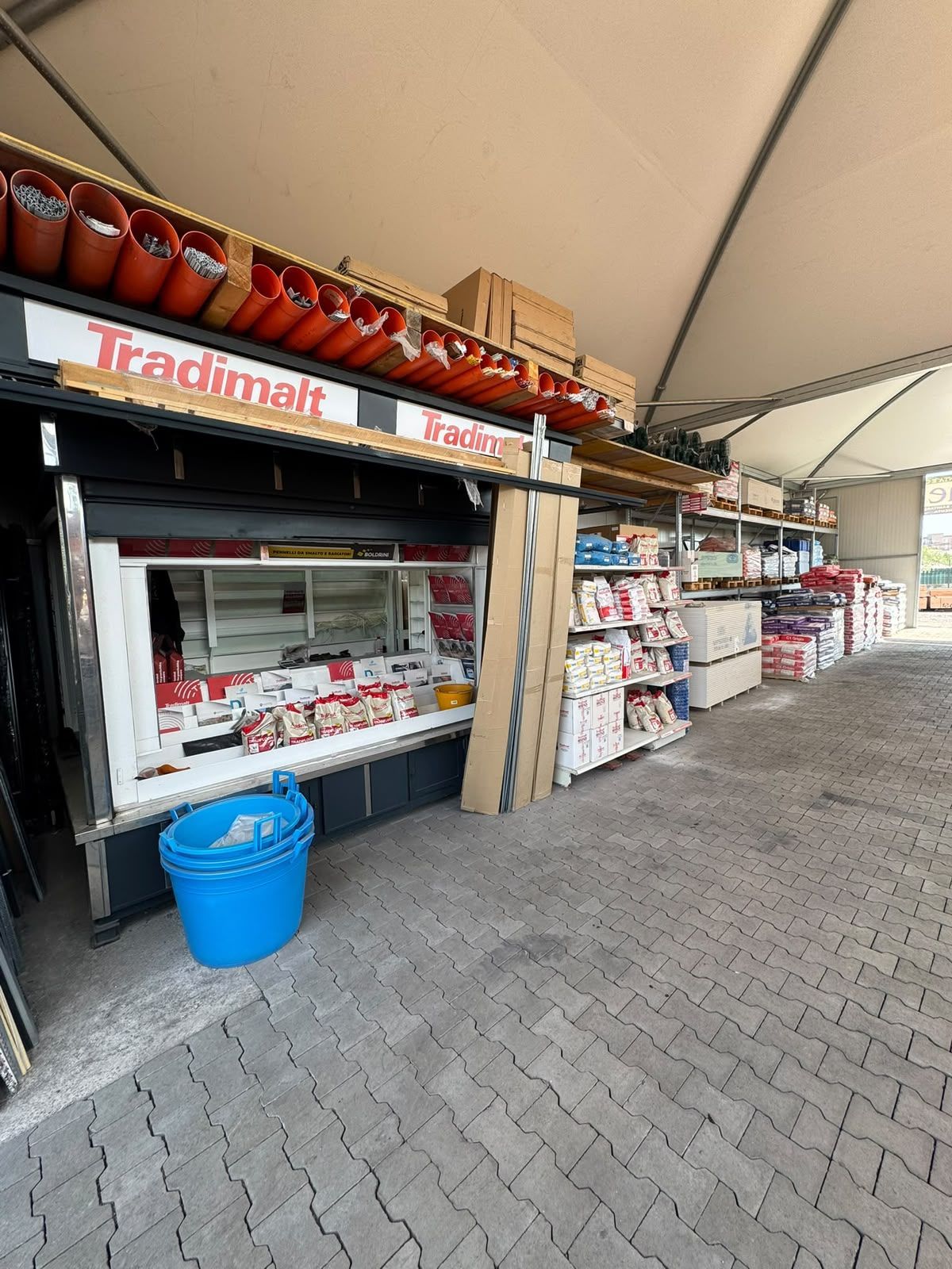 Bancarella di mercato all’aperto con insegna rossa, scaffali pieni di merce e un bidone blu sotto una tenda.