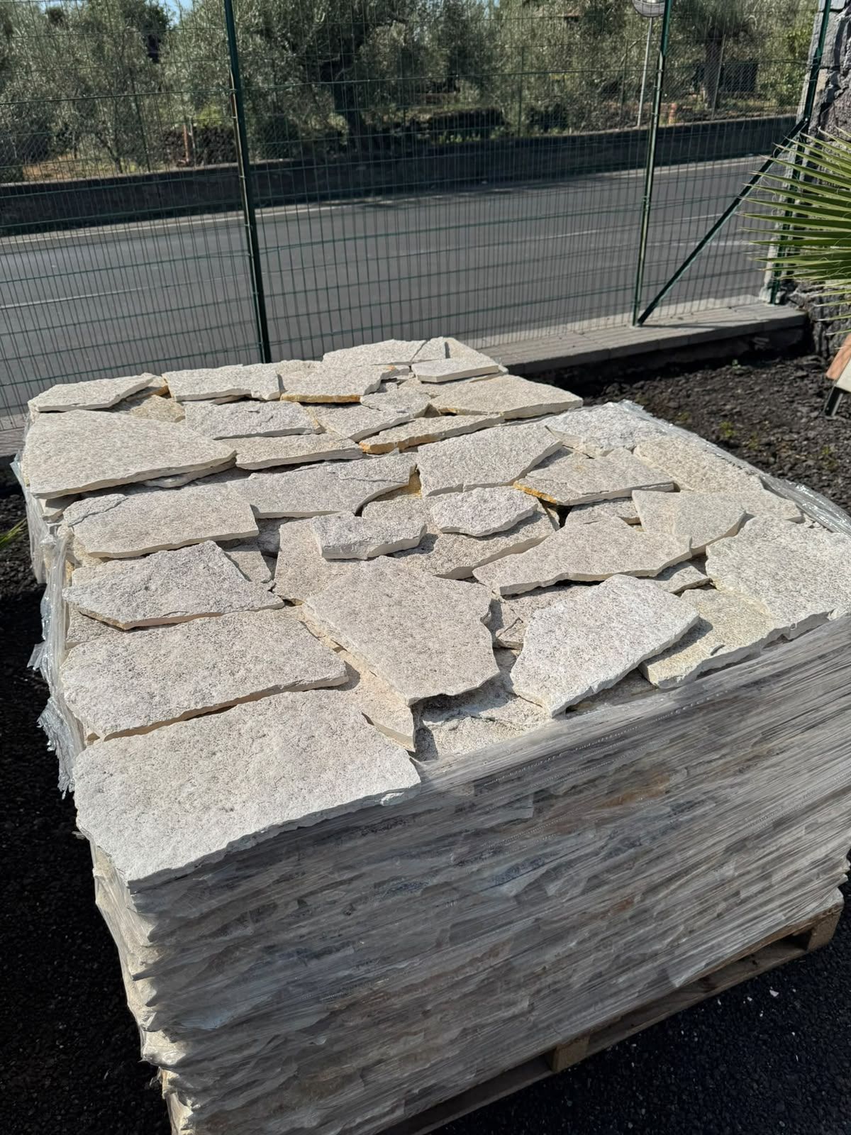 Pila di lastre di pietra chiara su un pallet all’aperto, vicino a una recinzione e a un terreno sterrato.