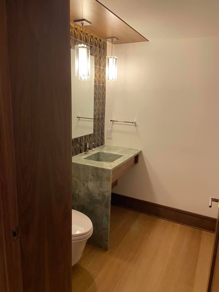 Small modern restroom with a concrete sink, mirror, pendant lights, and wooden floor; toilet partially visible.